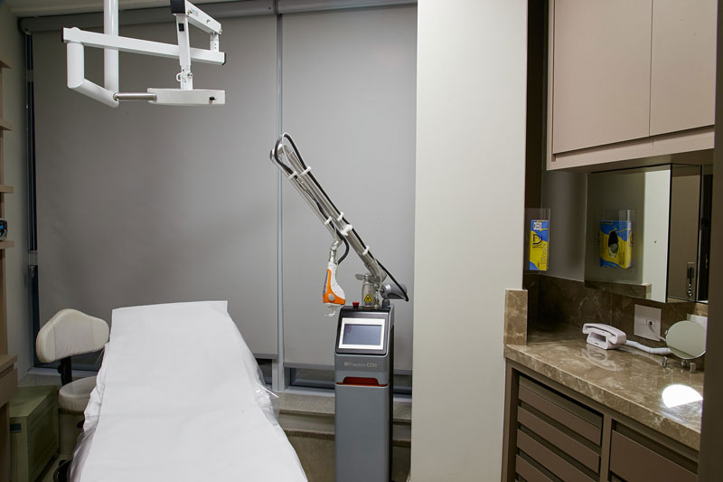 Sala de procedimento dermatológico com maca, aparelho de laser, bancada em mármore, armários e espelho.